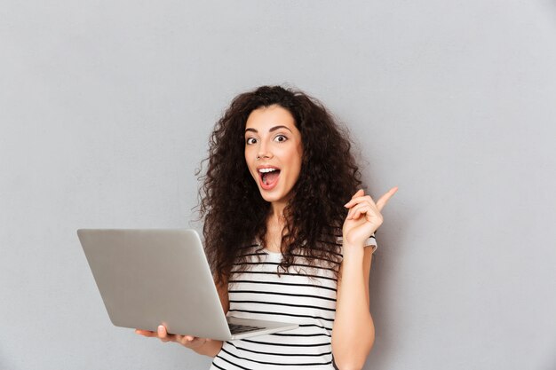 Portrait en studio de femme aux cheveux bouclés étant heureux de trouver des informations utiles sur Internet via un ordinateur argenté gesticulant eureka avec index