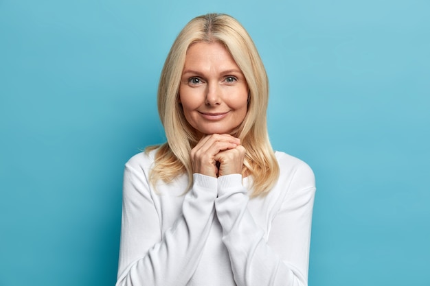 Portrait en studio d'une femme de cinquante ans confiante garde les mains sous le menton regarde directement la caméra avec une expression calme porte un pull blanc a bien soigné les poses de la peau