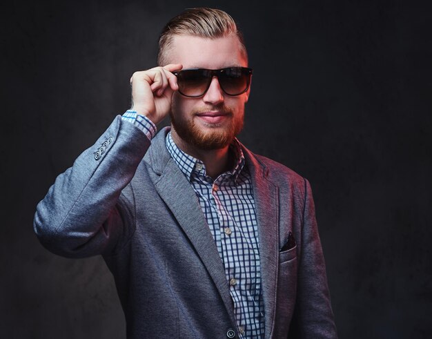 Portrait en studio d'un homme barbu rousse vêtu d'un costume et de lunettes de soleil sur fond gris.