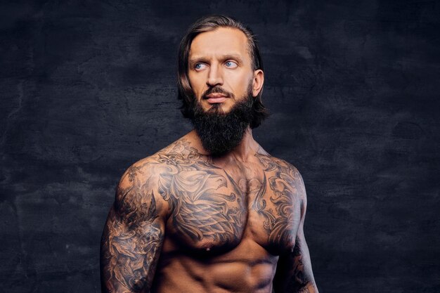 Portrait en studio d'un homme barbu tatoué et torse nu sur fond de vignette gris foncé.