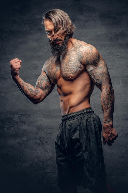 Portrait en studio d'un homme barbu tatoué et torse nu sur fond de vignette gris foncé.