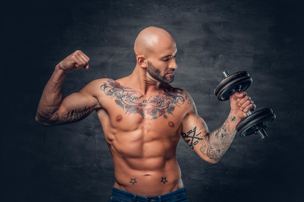 Portrait en studio d'un homme sportif à tête rasée avec des tatouages sur sa poitrine et ses bras tient une haltère.