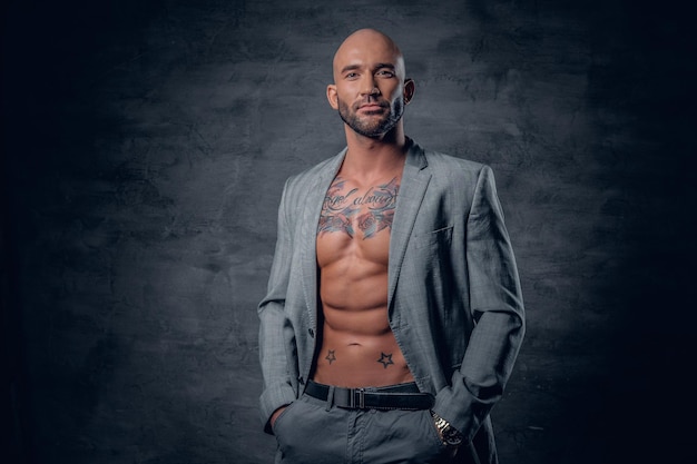 Portrait en studio d'un homme tatoué à la tête rasée vêtu d'une veste grise sur un torse nu.