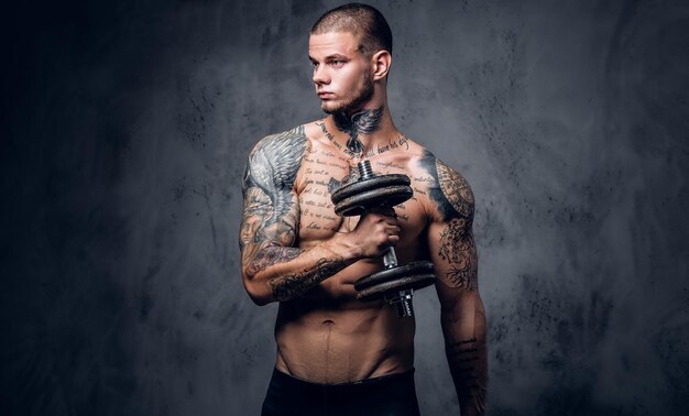 Portrait en studio d'un homme tatoué torse nu tenant une haltère sur un fond artistique gris.