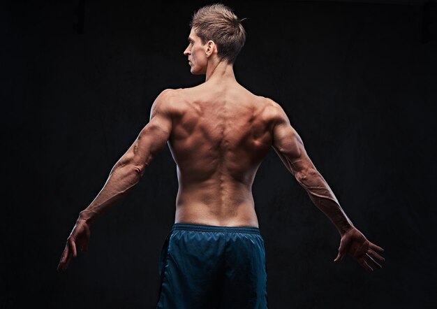 Portrait en studio d'un homme torse nu musculaire ectomorphe sur fond gris foncé. Vue arrière.