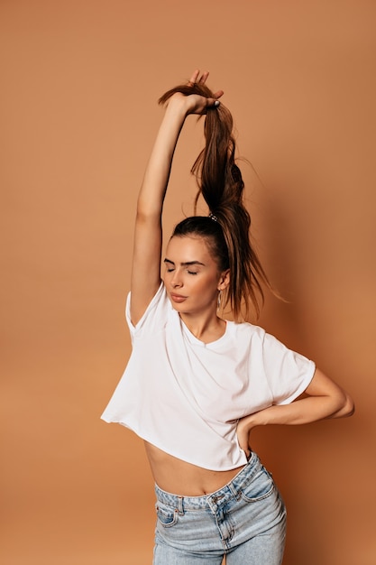 Portrait en studio intérieur de charmante jolie dame portant un t-shirt blanc et un jean posant sur un mur beige