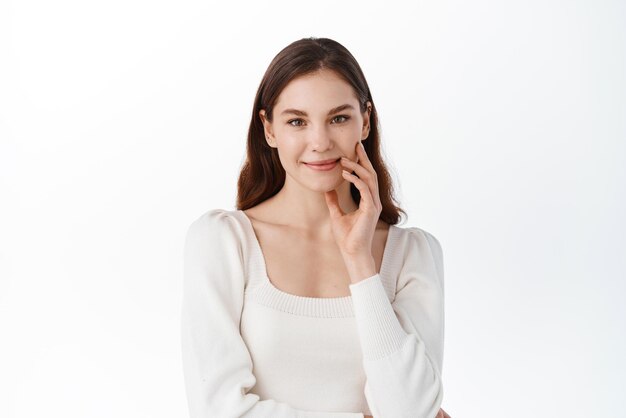 Portrait en studio de jeune femme montrant une émotion calme et détendue souriant doucement en regardant la caméra toucher le visage naturel avec un maquillage nu debout sur fond blanc