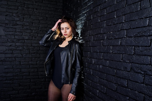 Portrait en studio de jeune fille brune sexy en veste de cuir noir contre le mur de briques