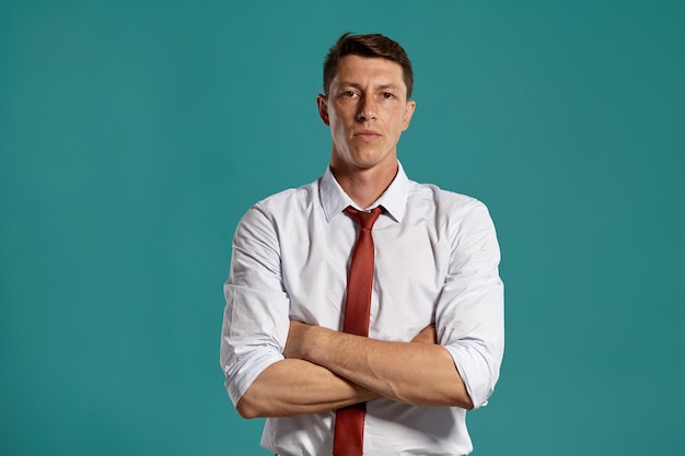 Portrait en studio d'un jeune homme de mauvaise humeur vêtu d'une chemise blanche classique et d'une cravate rouge croisant les mains et regardant la caméra tout en posant sur un fond bleu. Coupe de cheveux élégante. Concept d'émotions sincères. C