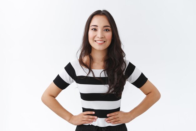 Portrait en studio jolie femme brune asiatique animée en t-shirt rayé tenant les mains sur les hanches debout confiant et optimiste souriant largement montrant une attitude déterminée et assurée