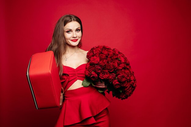 Portrait en studio à la mode d'une jeune femme élégante aux longs cheveux bruns en costume rouge cher avec un sac en cuir rouge et un bouquet de roses rouges Elle sourit à la caméra Lèvres rouges Fond rouge
