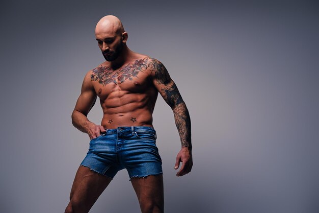 Portrait en studio de tête rasée torse nu, homme musclé avec des tatouages sur son torse vêtu d'un short en jean sur fond de vignette grise.