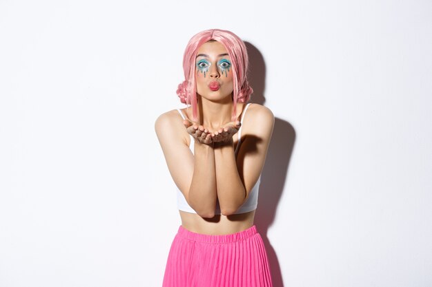 Portrait de stupide belle fille caucasienne avec un maquillage lumineux, habillée pour la fête en perruque rose glam, souffle de l'air baiser à la caméra, debout.