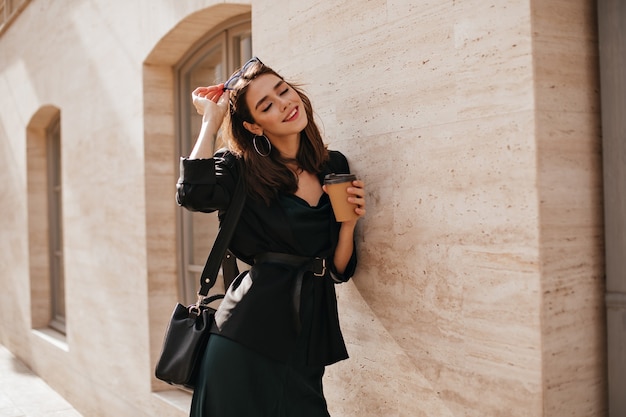 Portrait de style de vie d'une charmante jeune brune au maquillage lumineux, tenue sombre avec robe, veste, sac, lunettes de soleil et ceinture debout près d'un mur beige à l'extérieur et souriant