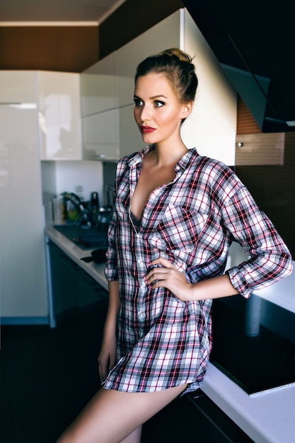 Portrait de style de vie intérieur de jolie femme blonde détendue et profiter de son heure du matin, posant à la cuisine, portant une chemise à carreaux confortable, des couleurs douces de film vintage.