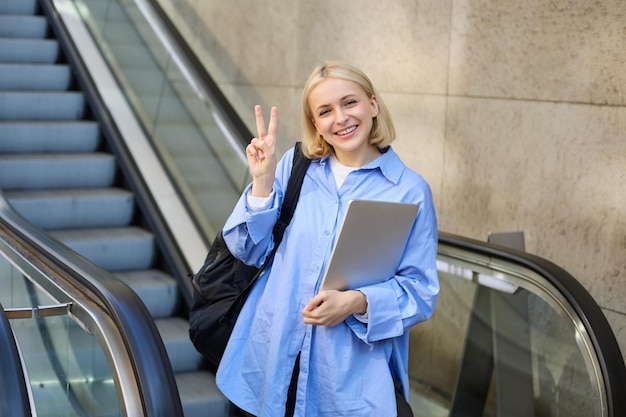 Portrait de style de vie d'une jeune femme positive étudiante avec un ordinateur portable et un sac à dos montrant la paix