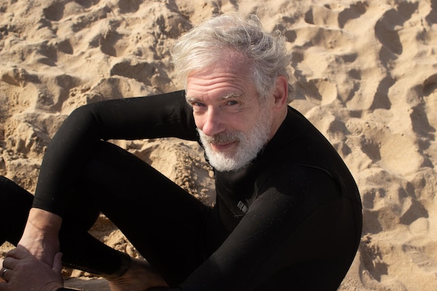 Photo gratuite portrait d'un surfeur masculin âgé reposant sur le sable. heureux homme barbu en combinaison assis sur la plage profitant du temps chaud de l'été, regardant la caméra. concept de sport actif et de repos pour la santé des personnes âgées