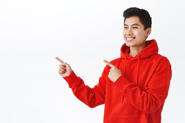 Portrait de taille d'un étudiant asiatique heureux et heureux en sweat à capuche rouge, boucle d'oreille, regardant et pointant les doigts avec une expression satisfaite, recommande un bon produit, mur blanc.