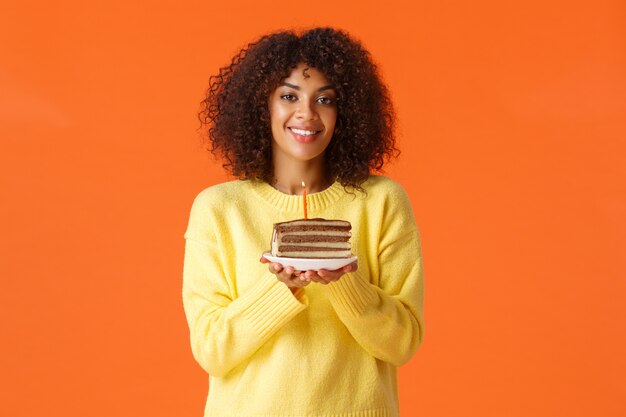 Portrait de taille fille afro-américaine de rêve b-day avec coupe de cheveux afro, tenant une assiette avec un gâteau d'anniversaire et une bougie allumée, soufflant pour faire un vœu, souriant joyeusement, célébrant sur un mur orange.