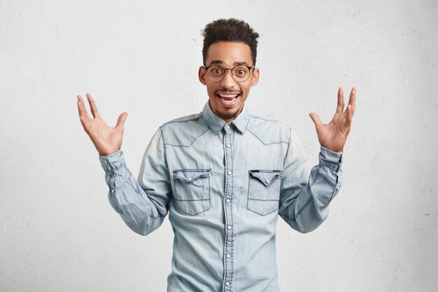 Portrait à la taille d'un homme à la mode ravi avec une coiffure à la mode, porte une chemise en jean, lève les mains