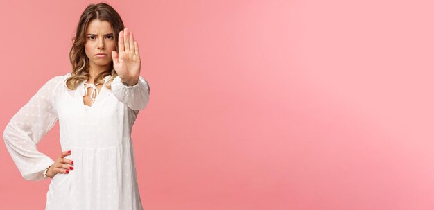 Portrait à la taille d'une jeune femme blonde sérieuse, affirmée et confiante, en robe blanche, étendre le bras en stop motion, fronçant les sourcils, demander d'arrêter d'interdire quelque chose d'avertissement exprimer un désaccord