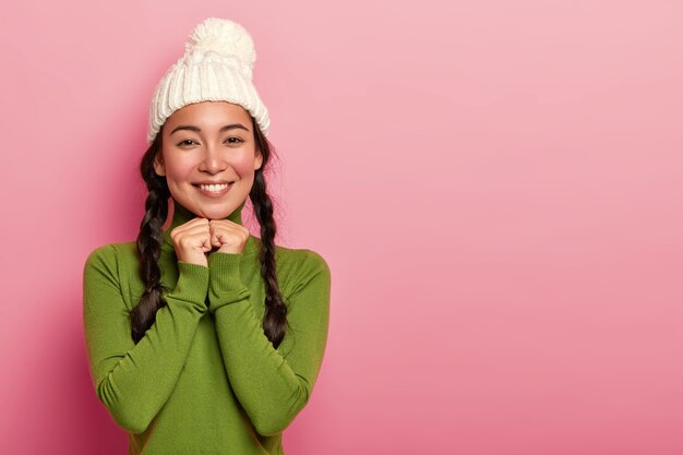 Portrait de tendre belle femme asiatique a les joues rouges, sourire agréable, garde les mains sous le menton, regarde heureusement directement à la caméra, porte un chapeau tricoté