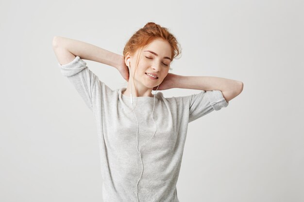 Portrait de tendre belle fille rousse, écouter de la musique dans les écouteurs avec les yeux fermés souriant en appréciant. Mains derrière la tête.