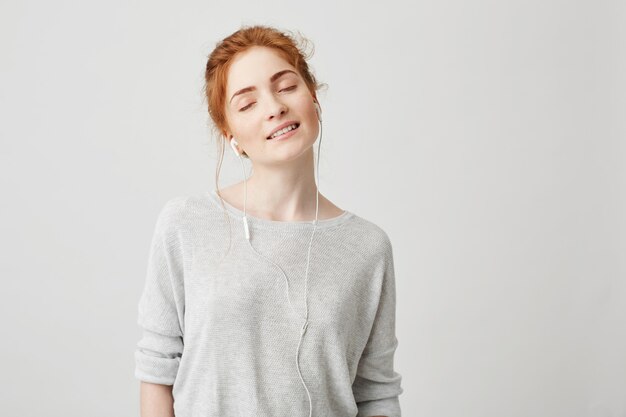 Portrait de tendre belle fille rousse, écouter de la musique dans les écouteurs avec les yeux fermés souriant en appréciant.