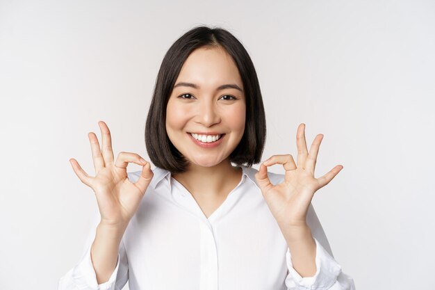 Portrait de tête en gros plan d'une fille asiatique montrant un signe correct ok et souriant satisfait recommandant d'être heureux de louer et de faire un compliment sur fond blanc