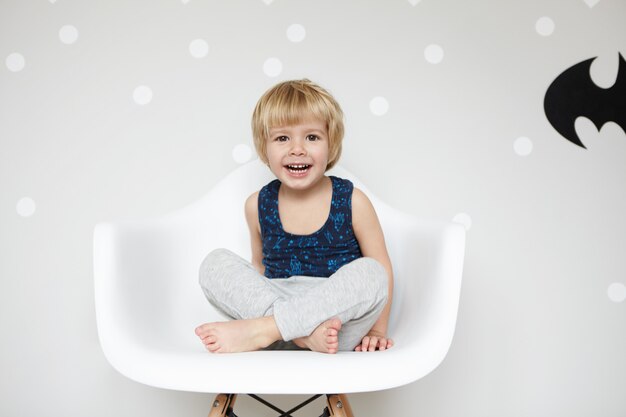 Portrait de tout-petit ludique aux cheveux blonds portant un pyjama, assis sur une chaise les jambes croisées, riant, bouche grande ouverte, montrant ses dents blanches, contre le mur blanc gagner