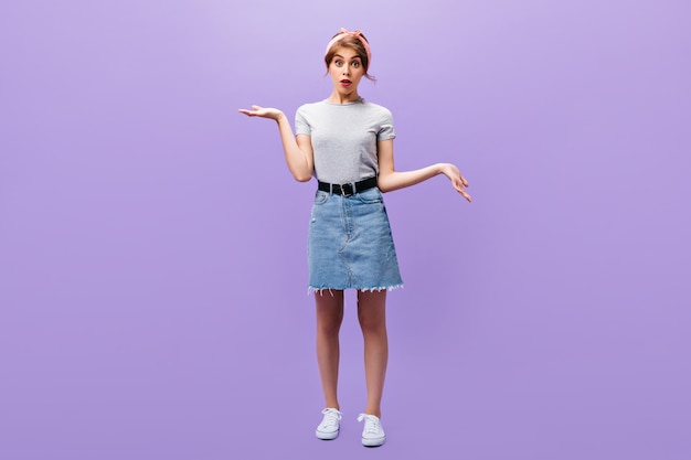 Portrait de toute la longueur de la jeune fille en jupe en jean et chemise grise. Femme surprise avec un bandeau rose dans des vêtements élégants et des baskets blanches posant.