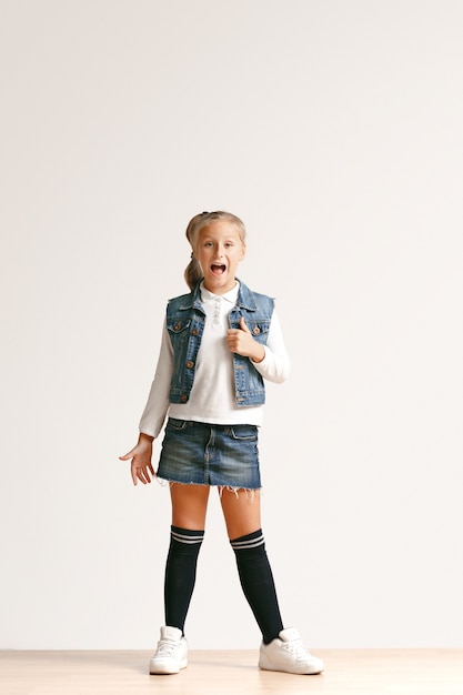 Portrait de toute la longueur de la mignonne petite adolescente dans des vêtements de jeans élégants regardant la caméra et souriant contre le mur blanc du studio. Concept de mode pour enfants