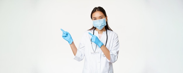 Portrait d'une travailleuse médicale asiatique pointant les doigts vers la gauche portant un masque facial et des gants en caoutchouc st