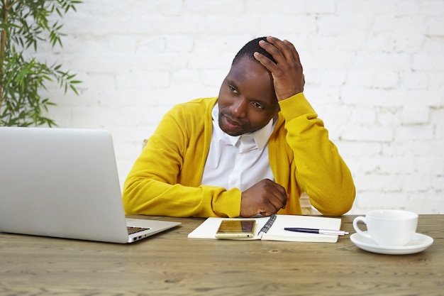 Portrait de triste employé afro-américain portant un cardigan jaune touchant la tête se sentant fatigué et surchargé de travail à cause du stress ou de l'échec au travail, assis au bureau avec ordinateur portable, café et agenda