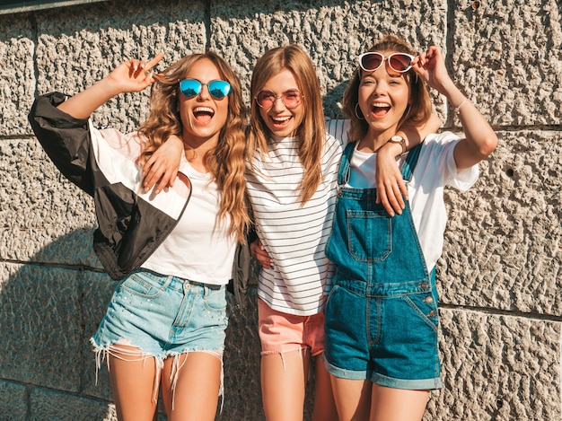 Portrait de trois jeunes belles filles hipster souriantes dans des vêtements d'été à la mode. Sexy, insouciant, femmes, poser, près, mur, rue, positif, modèles, amusant, lunettes soleil