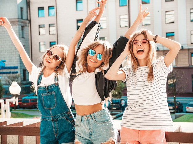 Portrait de trois jeunes belles filles hipster souriantes dans des vêtements d'été à la mode. Sexy, insouciant, femmes, poser, rue, positif, modèles, amusant, lunettes soleil, étreindre, élévation, mains