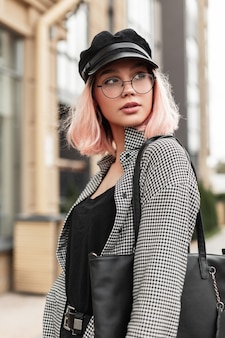 Portrait urbain d'une jeune femme élégante dans des lunettes vintage avec une coiffe dans une chemise à la mode avec un sac à main noir se promène dans la ville