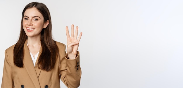 Portrait d'une vendeuse d'entreprise montrant le numéro quatre doigts et souriant debout en costume sur fond blanc