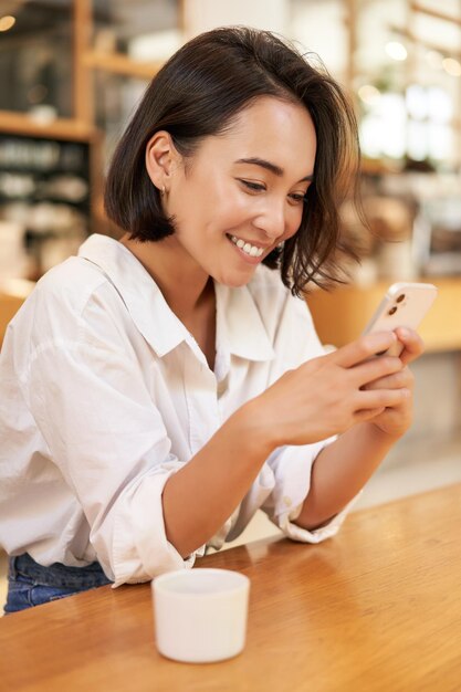 Portrait vertical d'une femme asiatique élégante assise dans un café, buvant du café et utilisant un smartphone