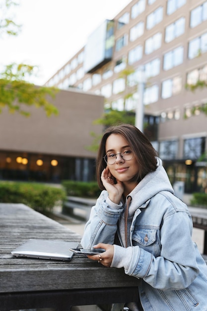 Photo gratuite portrait vertical d'une jeune étudiante séduisante assise sur le campus de l'université du parc près d'un point wifi étudiant en attente d'un ami utiliser un ordinateur portable et un téléphone portable souriant appareil photo joyeux