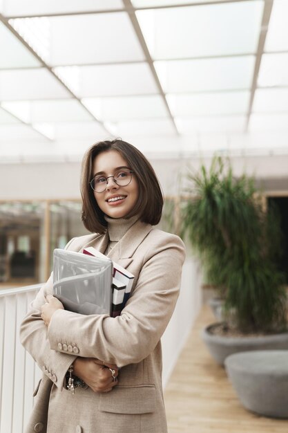 Portrait vertical d'une jeune femme employée ou entrepreneure réussie au tuteur de la salle de bureau portant un ordinateur portable et des cahiers en détournant les yeux avec un sourire satisfait et satisfait comme son travail