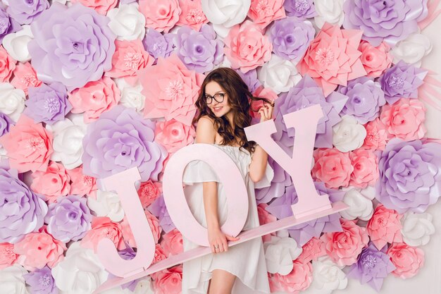 Portrait vertical de jolie fille brune. Elle se lève et tient le mot JOY en bois souriant largement. Elle a les cheveux bouclés et porte une robe blanche à l'épaule et des lunettes noires. Fond en fleurs roses.