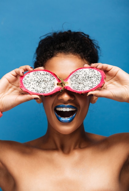 Portrait Vertical De Joyeuse Femme Afro-américaine Couchait Tout En Couvrant Les Yeux Avec Des Fruits Exotiques Pitahaya Coupés En Deux Isolé, Sur Bleu