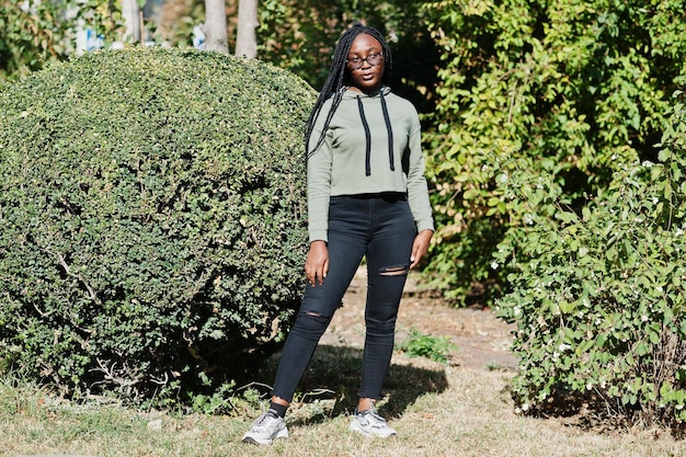 Photo gratuite portrait de la ville d'une jeune femme positive à la peau foncée portant un sweat à capuche vert et des lunettes contre la verdure