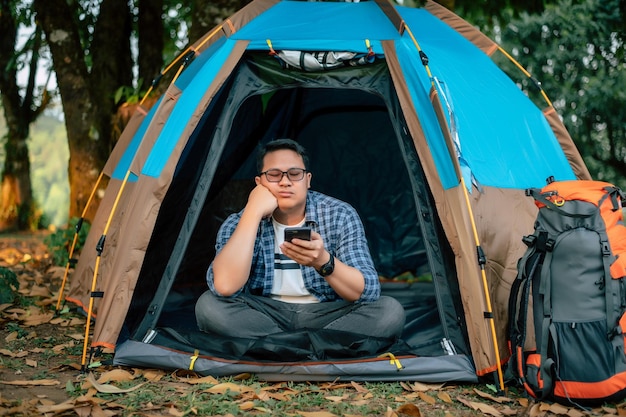 Portrait d'un voyageur asiatique s'ennuyant avec des lunettes à l'aide d'un smartphone dans une tente de camping