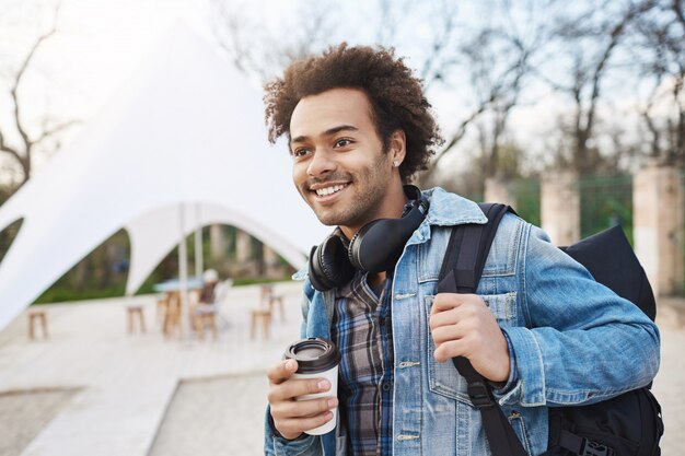 Portrait de vue latérale d'un mec à la peau sombre heureux à la mode tenant un sac à dos et du café en marchant dans la ville, souriant et regardant de côté, étant de bonne humeur