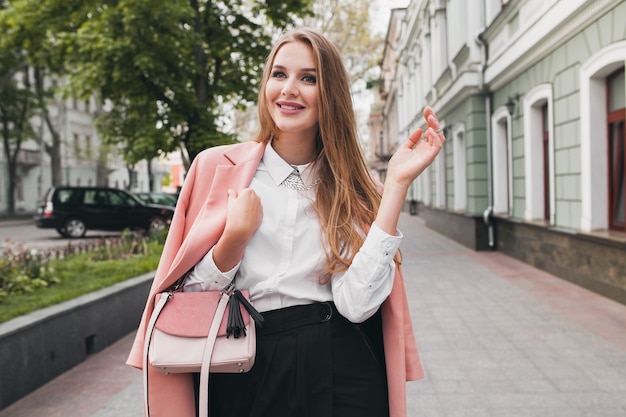 Posant une jolie femme souriante élégante qui marche rue de la ville en manteau rose tendance de la mode printemps, style élégant