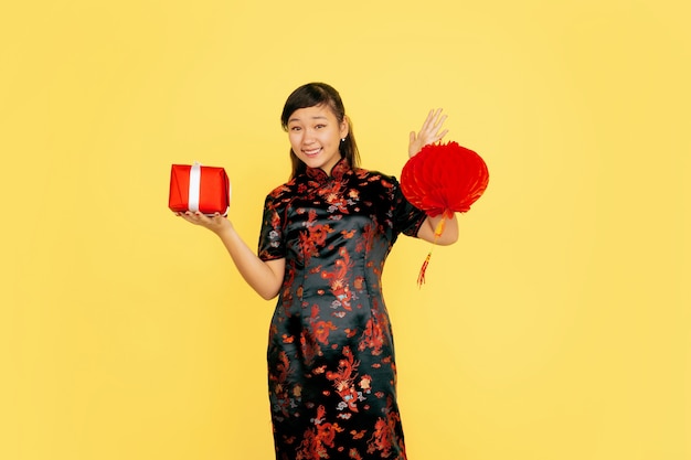 Posant avec lanterne et cadeau, souriant. Joyeux nouvel an chinois 2020. Portrait de jeune fille asiatique sur fond jaune. Le modèle féminin en vêtements traditionnels a l'air heureux. Célébration, émotions. Copyspace.