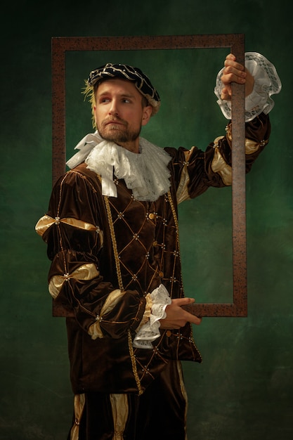 Photo gratuite posant réfléchie. portrait de jeune homme médiéval en vêtements vintage avec cadre en bois sur fond sombre. modèle masculin en tant que duc, prince, personne royale. concept de comparaison des époques, moderne, mode.