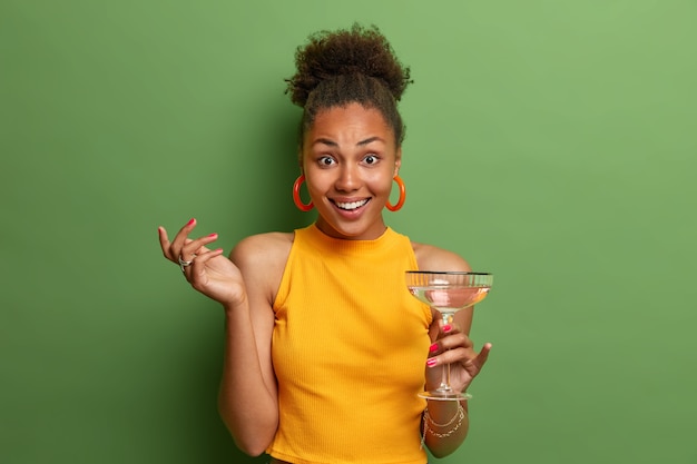 Positif, bonne pose féminine ethnique avec un verre de cocktail d'été, a l'air heureux et une conversation agréable avec un ami, porte des vêtements jaunes isolés sur un mur vert. Gens, boire, concept de loisirs